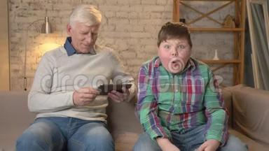 祖父和孙子坐在沙发上。 一个老人用平板电脑，一个年轻的胖子在游戏机上玩